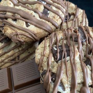 May include: Close-up of several chocolate chip cookies drizzled with melted chocolate. The cookies have large chocolate chunks and are sitting next to a bar of chocolate. The cookies are a light brown color, and the chocolate drizzle is a darker brown.
