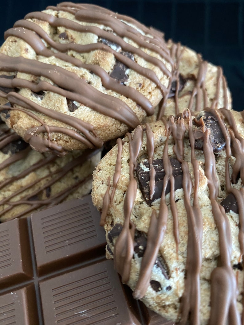 May include: Close-up of several chocolate chip cookies drizzled with melted chocolate. The cookies have large chocolate chunks and are sitting next to a bar of chocolate. The cookies are a light brown color, and the chocolate drizzle is a darker brown.