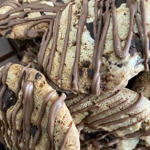 May include: Close-up of several chocolate chip cookies drizzled with melted chocolate. The cookies are golden brown with visible chocolate chips and a soft texture. Some chocolate bars are in the background.
