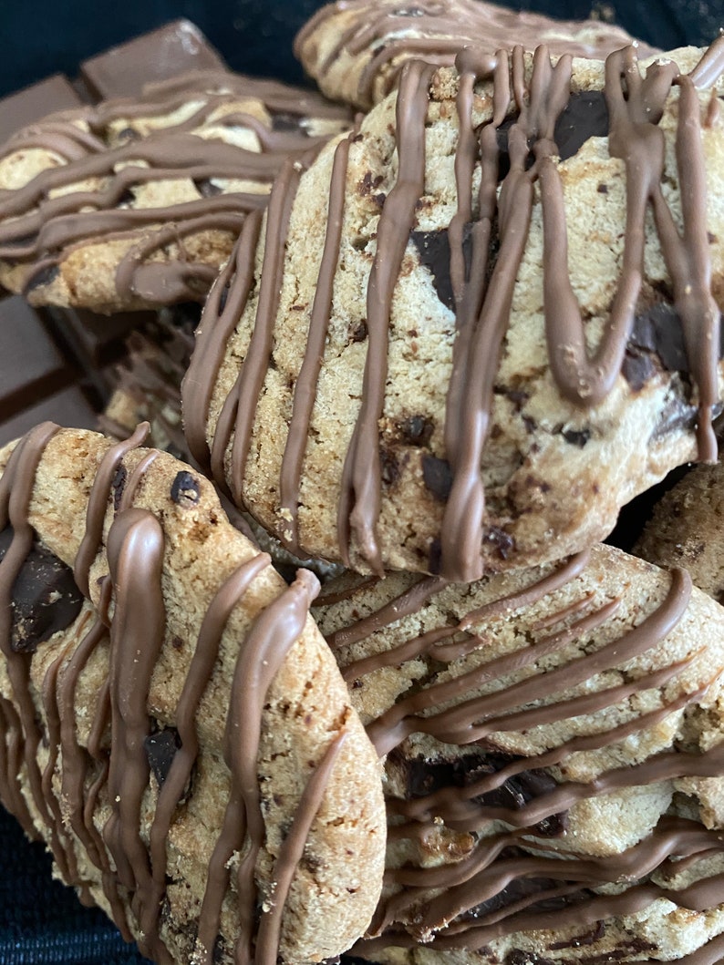 May include: Close-up of several chocolate chip cookies drizzled with melted chocolate. The cookies are golden brown with visible chocolate chips and a soft texture. Some chocolate bars are in the background.