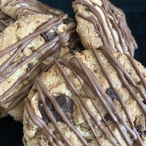 May include: Close-up of several chocolate chip cookies drizzled with melted chocolate. The cookies have a golden-brown color with visible chocolate chunks. The dark chocolate drizzle adds a contrasting color and texture to the baked goods.