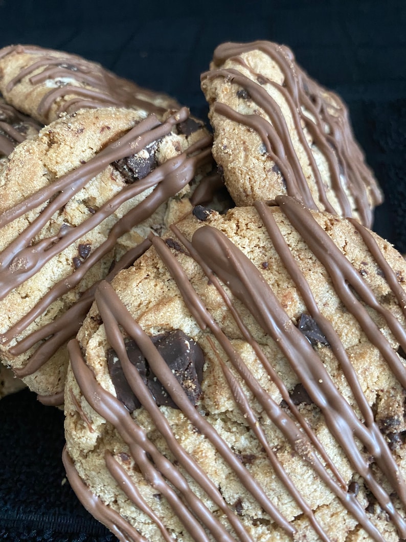 May include: Close-up of several chocolate chip cookies drizzled with melted chocolate. The cookies have a golden-brown color with visible chocolate chunks. The dark chocolate drizzle adds a contrasting color and texture to the baked goods.