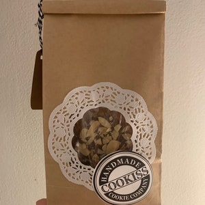 May include: A brown paper bag with a clear window revealing cookies. The bag has a white doily and a round sticker that reads "HANDMADE COOKISS COOKIE COMPANY". A brown tag hangs from the top with black and white twine.