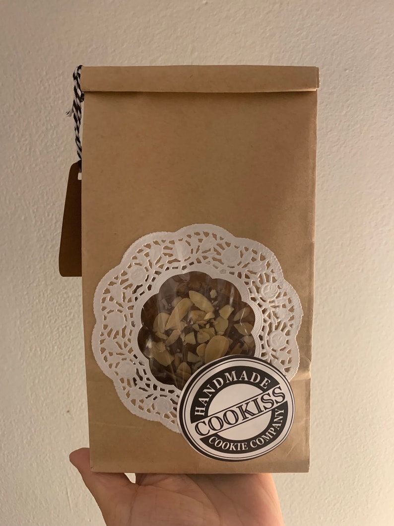 May include: A brown paper bag with a clear window revealing cookies. The bag has a white doily and a round sticker that reads "HANDMADE COOKISS COOKIE COMPANY". A brown tag hangs from the top with black and white twine.
