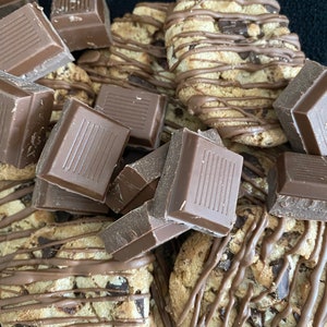 May include: Close-up of chocolate chip cookies drizzled with chocolate and surrounded by chunks of chocolate bars. The cookies have a golden-brown color with visible chocolate chips.