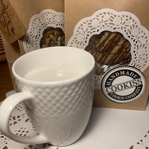 May include: A white ceramic mug with a textured diamond pattern sits on a white paper doily. Behind the mug are two brown paper bags with cookies visible through a clear window, and a "Handmade Cookiss Cookie Company" sticker.