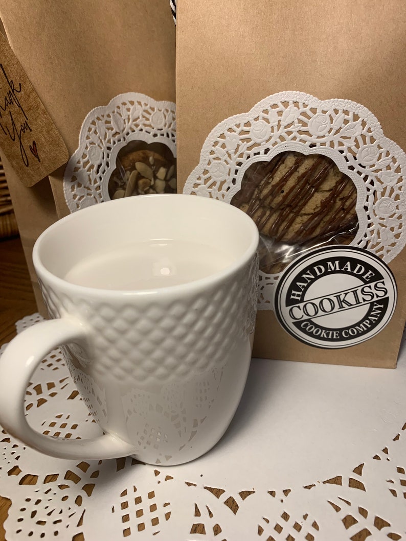 May include: A white ceramic mug with a textured diamond pattern sits on a white paper doily. Behind the mug are two brown paper bags with cookies visible through a clear window, and a "Handmade Cookiss Cookie Company" sticker.