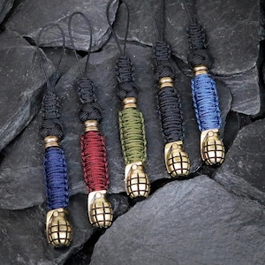 May include: Five lanyard charms featuring a brass-toned metal grenade-shaped bead at the base, attached to a braided paracord section in blue, red, olive green, black, and blue, finished with a black cord loop for attachment to various items.