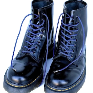 Könnte beinhalten: Ein Paar schwarze Lederstiefel mit blauen Schnürsenkeln. Die Stiefel haben ein gelbes Etikett mit der Aufschrift "AirWair" an der Seite.