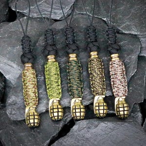 May include: Five paracord lanyards featuring brass-colored metal grenade charms at the base. Each lanyard has a black braided cord loop at the top, a brass-colored metal connector, and a woven paracord handle in various camouflage patterns.