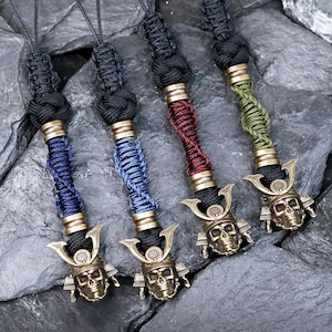 May include: Four keychains with a samurai skull design. Each keychain features a black lanyard, a brass-colored metal accent, and a skull pendant. The lanyards are wrapped in blue, red, and green threads. The skull pendants have a helmet design.