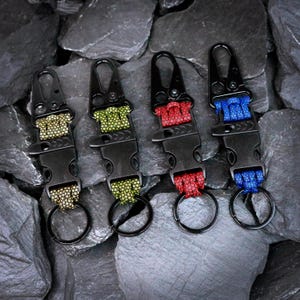May include: Four tactical keychains featuring black plastic side-release buckles and metal carabiner clips. Each keychain includes a woven paracord section in tan, olive green, red, or blue, finished with a circular metal split ring at the base.