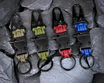 Puede incluir: Cuatro llaveros tácticos con hebillas de liberación lateral de plástico negro y mosquetones de metal. Cada llavero incluye una sección de paracord trenzado en color beige, verde oliva, rojo o azul, con una anilla metálica circular.