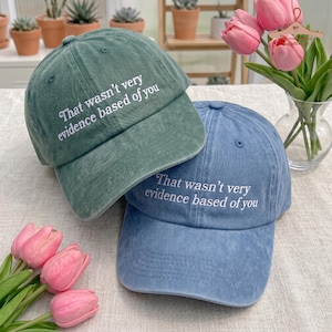 May include: A sage green baseball cap and a denim blue baseball cap, both featuring the white embroidered text "That wasn't very evidence based of you" on the front panels. Each cap has a structured crown, a curved brim, and a washed fabric finish.