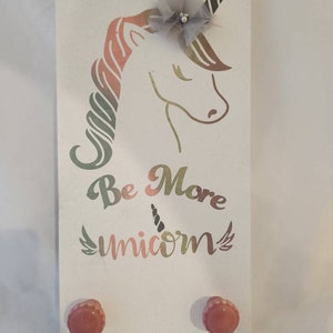 May include: White wooden wall decor featuring a unicorn design with a gray horn and a decorative bow. The text "Be More Unicorn" is printed below the unicorn. Two red knobs are at the bottom.