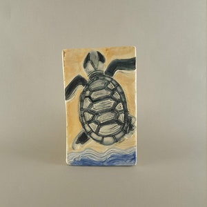 Puede incluir: Un azulejo de cerámica con una imagen pintada a mano de una tortuga marina nadando en el océano. La tortuga es gris y blanca, y el océano es azul y blanco.
