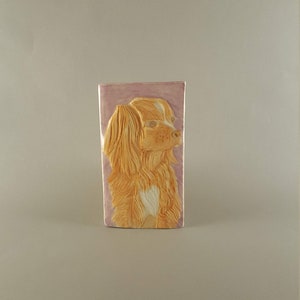 Puede incluir: Un azulejo de cerámica con una escultura en relieve de la cabeza de un perro. El perro es de color marrón claro con marcas blancas en el pecho y un fondo rosa.