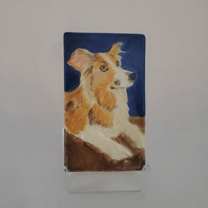 May include: A hand-painted ceramic tile featuring a portrait of a brown and white dog with a blue background.