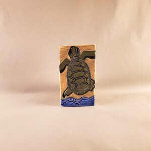 Puede incluir: Un azulejo de cerámica con una tortuga marina verde nadando en agua azul. La tortuga está mirando hacia la derecha y tiene un caparazón detallado. El azulejo es rectangular y tiene un fondo marrón claro.