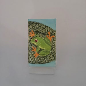 May include: A green tree frog with orange feet is perched on a green leaf with a blue background. The frog has red eyes.