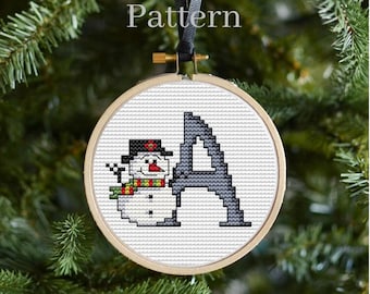 Könnte beinhalten: Runder Kreuzstich-Weihnachtsschmuck mit einem Schneemann und dem Buchstaben "A". Der Schneemann trägt einen schwarzen Hut und einen bunten Schal. Der Schmuck befindet sich in einem Holzreifen und hat ein schwarzes Band zum Aufhängen. Das Wort "Pattern" steht oben.