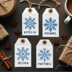 Könnte beinhalten: Vier weiße Geschenkanhänger mit blauen Kreuzstich-Schneeflockenmustern und den Namen Amelia, Michael, Jessica und Matthew. Jeder Anhänger hat eine braune Öse und ist mit Bindfaden versehen. Geschenke und Gewürze sind um die Anhänger angeordnet.