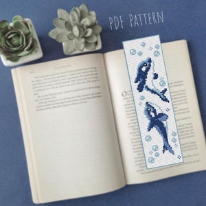 Könnte beinhalten: Ein PDF-Muster für ein Kreuzstich-Lesezeichen mit drei blauen Delfinen, die auf einem weißen Hintergrund mit blauen Blasen schwimmen. Der Text "PDF PATTERN" befindet sich oben im Bild.