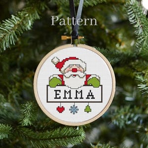 May include: A cross-stitch ornament featuring a cartoon Santa Claus holding a sign with the name "EMMA" on it. The ornament is decorated with a red heart, a snowflake, and a green Christmas tree.