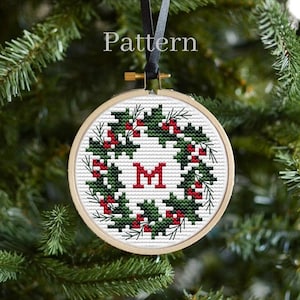 May include: A cross-stitch pattern for a Christmas bauble featuring a holly wreath with red berries and a white letter "M" in the centre. The pattern is displayed in a wooden embroidery hoop.