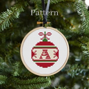 May include: A cross-stitch Christmas ornament in a wooden hoop, featuring a red and white design with the letter "A". The ornament is hanging from a black ribbon against a backdrop of green Christmas tree branches. The word "Pattern" is at the top.