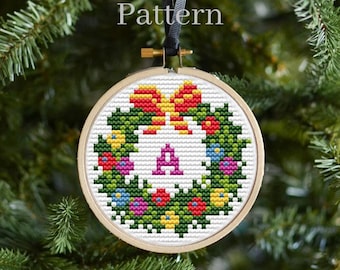 Puede incluir: Un patrón de punto de cruz para un adorno navideño con una corona con un lazo rojo y la letra A en el centro. La corona está decorada con flores de colores y hojas verdes.