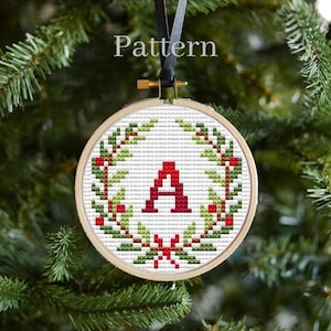 May include: A cross-stitch ornament featuring a green wreath with red berries and a red letter "A" in the center. The ornament is hanging on a Christmas tree.