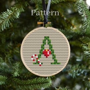 Könnte beinhalten: Ein Kreuzstich-Weihnachtsschmuck mit dem Buchstaben "A" in Grün, einem roten Herzen und Zuckerstangen. Der Schmuck befindet sich in einem Holzreifen und hängt an einem schwarzen Band. Das Wort "Pattern" ist oben aufgedruckt.