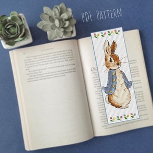 May include: A PDF pattern for cross-stitching a bookmark featuring Peter Rabbit in a blue jacket. The bookmark is surrounded by small yellow, red, and green flowers.