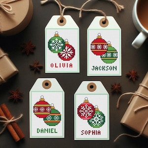 May include: Four Christmas gift tags with cross-stitched ornament designs in red and green. Each tag has a name: Olivia, Jackson, Daniel, and Sophia. The tags are on a dark surface with gifts and spices.
