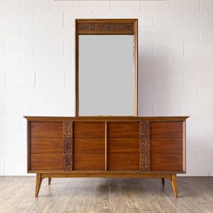 May include: A mid-century modern dresser and mirror set. The dresser is made of wood with a warm brown finish, featuring six drawers and decorative carvings. The mirror is rectangular with a wooden frame.