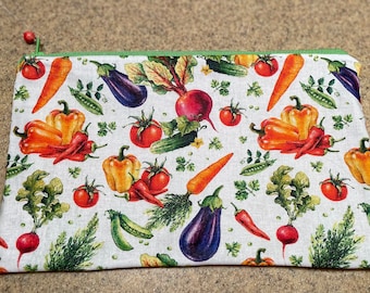 Könnte beinhalten: Eine weiße Reißverschlusstasche mit einem grünen Reißverschluss. Die Tasche ist mit einem bunten Muster aus Gemüse verziert, darunter Karotten, Paprika, Auberginen, Tomaten und Radieschen.