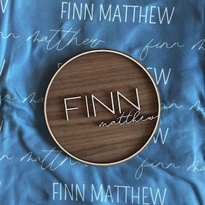 Op de afbeelding: Een rond houten bord met de naam "FINN matthew" gegraveerd in witte letters. Het bord bevindt zich op een blauwe stoffen achtergrond met dezelfde naam die in een cursief schrift wordt herhaald.