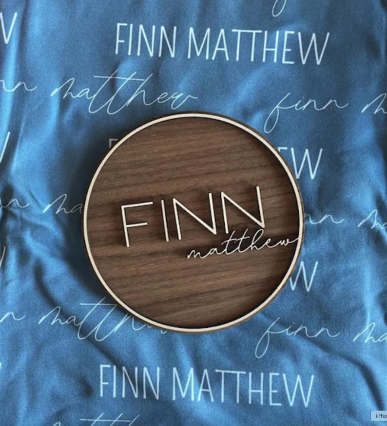 Op de afbeelding: Een rond houten bord met de naam "FINN matthew" gegraveerd in witte letters. Het bord bevindt zich op een blauwe stoffen achtergrond met dezelfde naam die in een cursief schrift wordt herhaald.