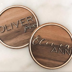 May include: Two round wooden signs with names written in white script. The first sign says "Oliver John" and the second sign says "Olivia Ann".