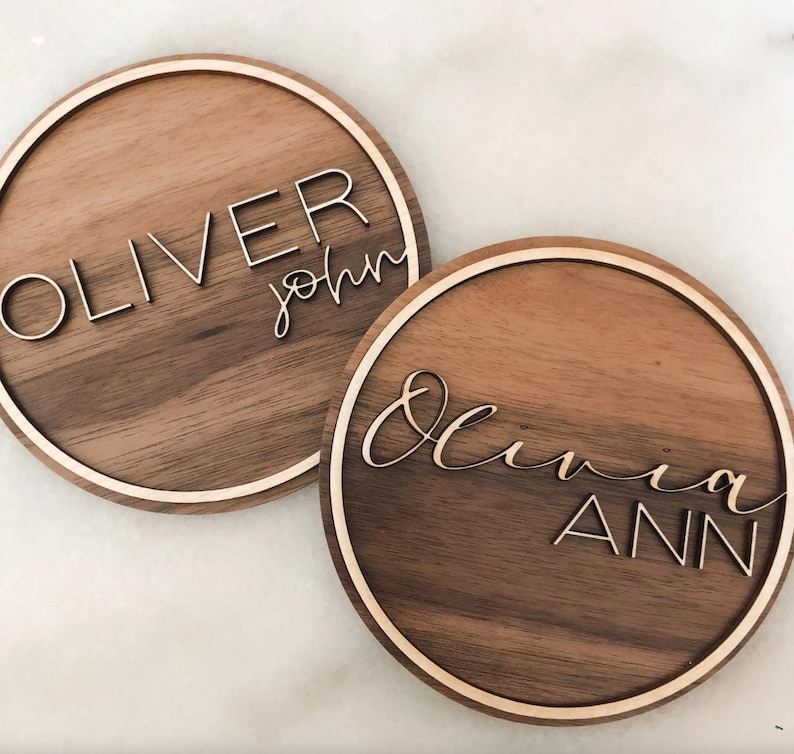 Op de afbeelding: Twee ronde houten borden met namen in witte letters. Het eerste bord zegt "Oliver John" en het tweede bord zegt "Olivia Ann".