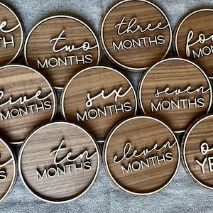 May include: A set of twelve wooden milestone discs. Each disc is round with a light wood border and a darker wood center. White script lettering indicates months one through twelve, and one year.