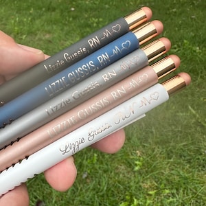 May include: Five personalized pens with rose gold accents. The pens are engraved with the name "Lizzie Gussis" and the title "RN-M". The pens are in various colors, including gray, blue, silver, pink, and white.