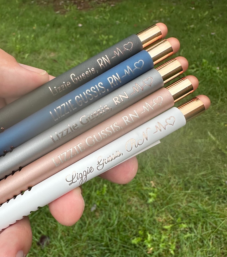 May include: Close-up view of six engraved pens in varying pastel colors: gray, blue, silver, rose gold, and white. Each pen features the same engraved text: 'Lizzie Gussis, RN-♡' with a heart and EKG symbol. The pens have rose gold colored accents on the top.