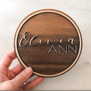 Op de afbeelding: Een rond houten bord met een lichtbruine houtnerf en een lichtbruine buitenring. Het bord heeft de naam "Olivia Ann" gegraveerd in een cursief schrift.