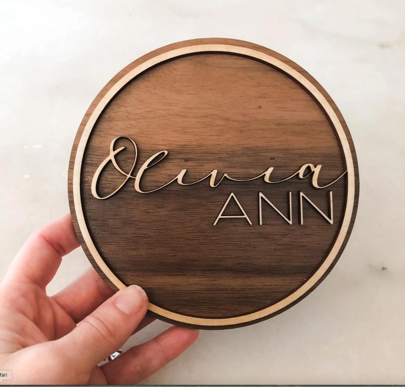 Op de afbeelding: Een rond houten bord met een lichtbruine houtnerf en een lichtbruine buitenring. Het bord heeft de naam "Olivia Ann" gegraveerd in een cursief schrift.
