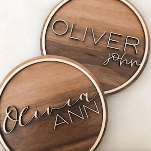Op de afbeelding: Twee ronde houten borden met namen in cursief schrift. Het bovenste bord zegt "Oliver John" en het onderste bord zegt "Olivia Ann".