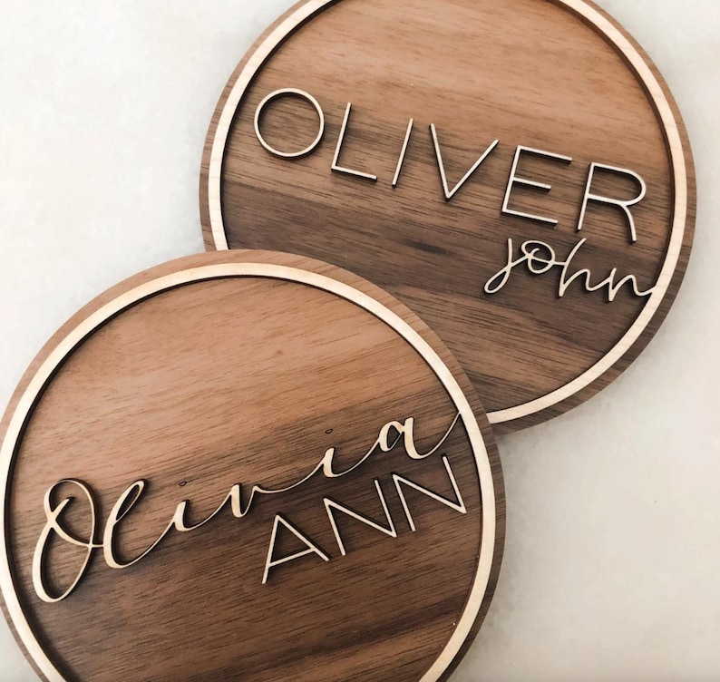 Op de afbeelding: Twee ronde houten borden met namen in cursief schrift. Het bovenste bord zegt "Oliver John" en het onderste bord zegt "Olivia Ann".