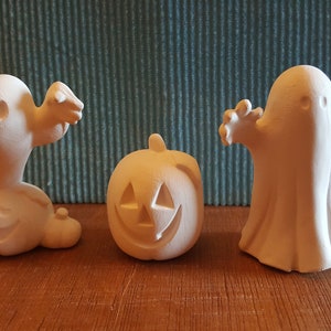 May include: Three unpainted ceramic Halloween decorations: two ghosts and a carved pumpkin. The ghost figures have outstretched arms, and the pumpkin has a carved face. The decorations are on a wooden surface.