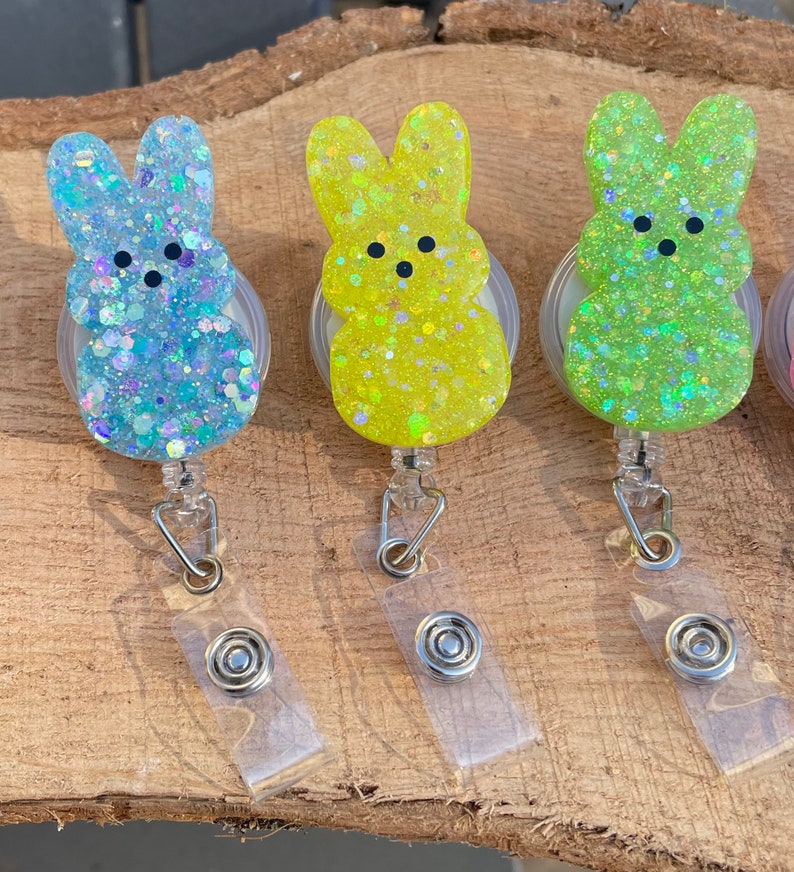 Op de afbeelding: Drie pastelkleurige, glinsterende, intrekbare badgehouders in de vorm van een konijn. De konijnenvormen zijn lichtblauw, geel en groen.